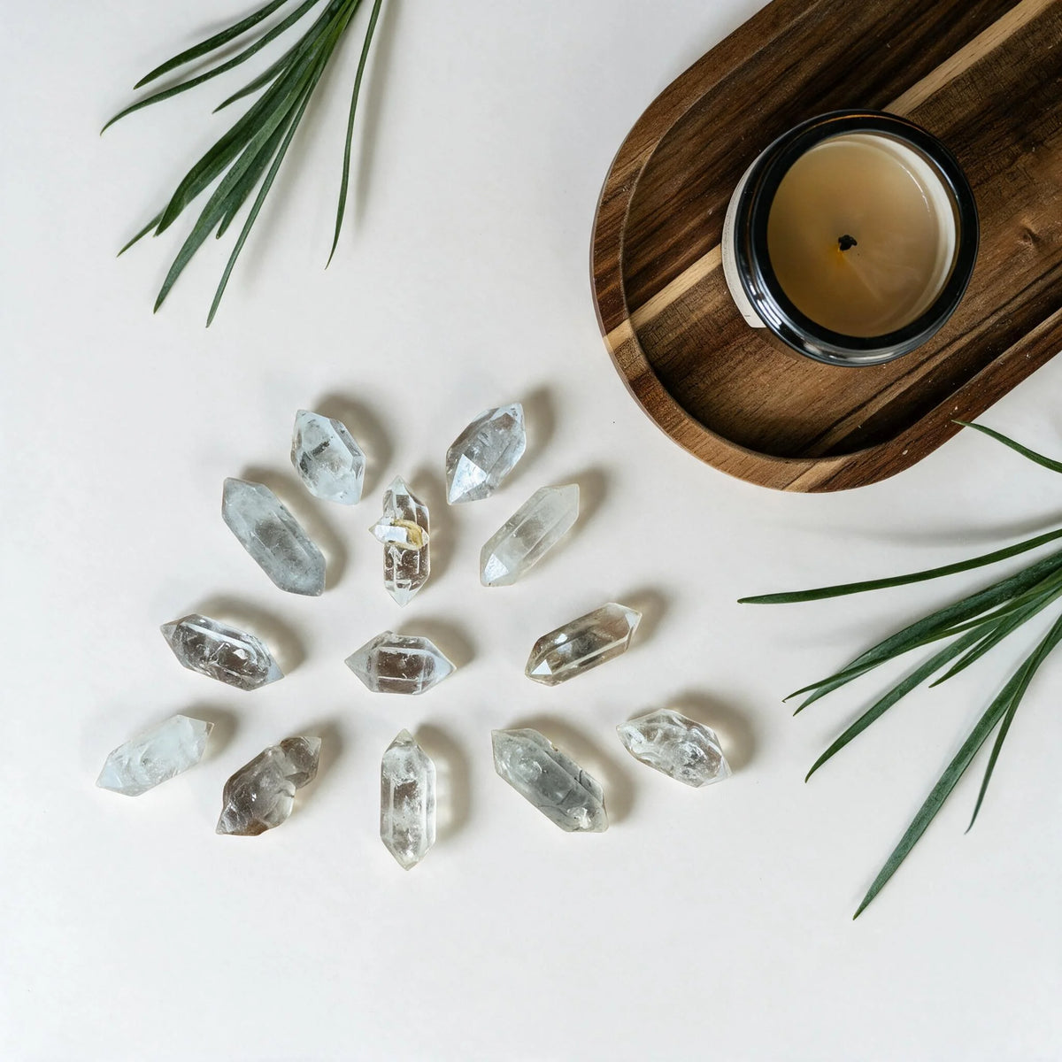 A circular grid arrangement of natural Ember Quiet Twin Peak clear quartz crystals on a bright, minimalist aesthetic background. Accompanied by a wooden tray, a relaxing candle, and fresh green leaves, creating a calm and healing energy space.