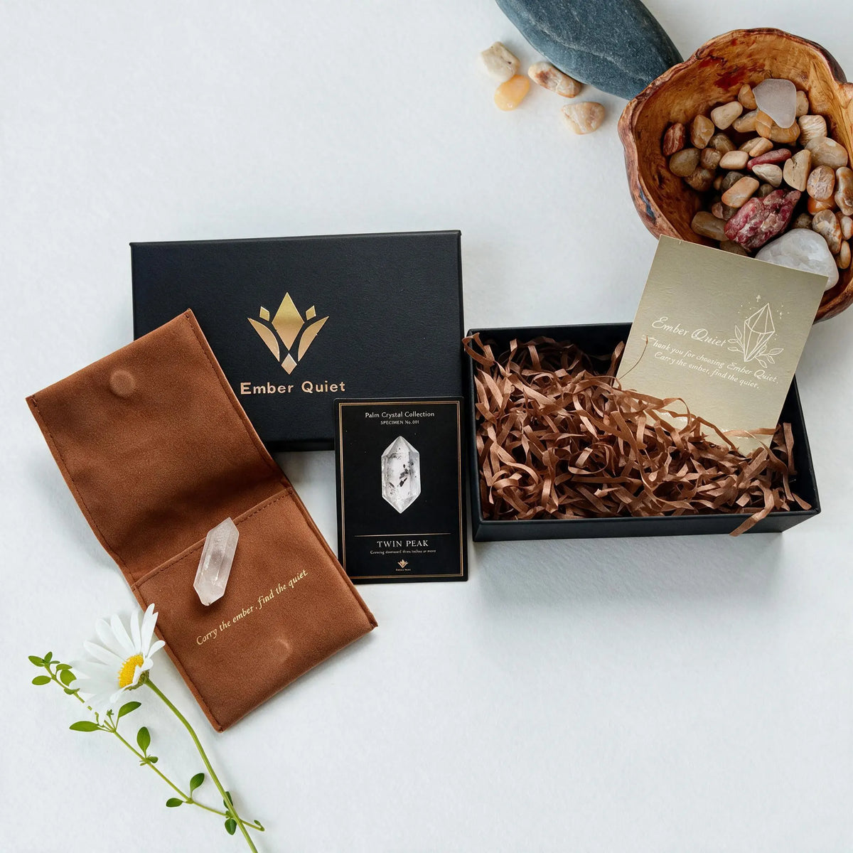 Ember Quiet premium crystal gift box packaging. A black unboxing experience featuring a gold logo, brown velvet pouches, an elegant information card, and a raw clear quartz crystal resting on a natural beige surface.